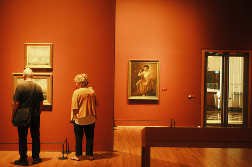 Two people in a museum observing paintings on an orange wall.